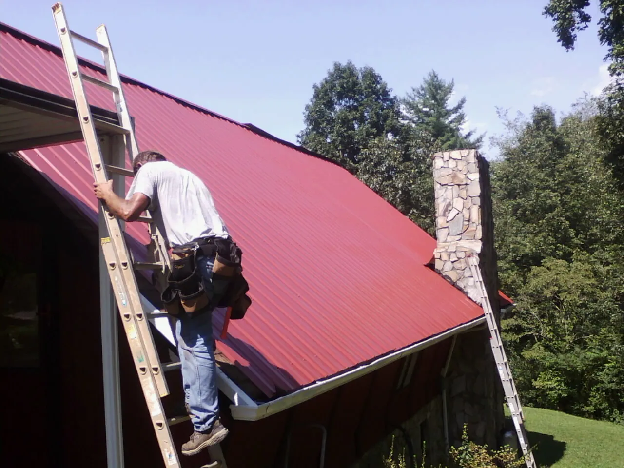 Roofer on a ladder beside a red metal roof and stone chimney — Soffit & Fascia Repair by Roofing Brothers in Worthington Springs